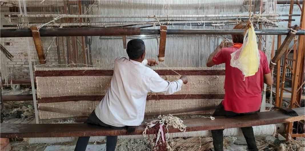 Two weavers seated at a pit loom in Bhadohi, working a striped flatweave across the warp